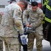 Several U.S. Army soldiers in OCP (Operational Camouflage Pattern) uniforms are gathered near a fire truck, examining or handling a medical/electronic device. One soldier, identified by name tape as 'OSMA,' is actively handling what appears to be a portable medical monitor or defibrillator. A firefighter in high-visibility gear is visible on the right side of the frame, suggesting an interagency training or coordination exercise.