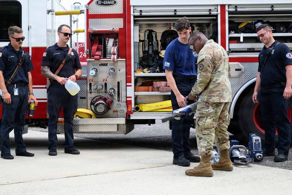 A group of firefighters and a military soldier in Army Combat Uniform (ACU/OCP camouflage) are gathered behind an E-ONE fire truck, appearing to conduct a demonstration or equipment exchange. The soldier is handling what appears to be a tool or piece of equipment while firefighters observe. The setting appears to be a fire station or emergency services facility.