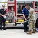 A group of firefighters and a military soldier in Army Combat Uniform (ACU/OCP camouflage) are gathered behind an E-ONE fire truck, appearing to conduct a demonstration or equipment exchange. The soldier is handling what appears to be a tool or piece of equipment while firefighters observe. The setting appears to be a fire station or emergency services facility.