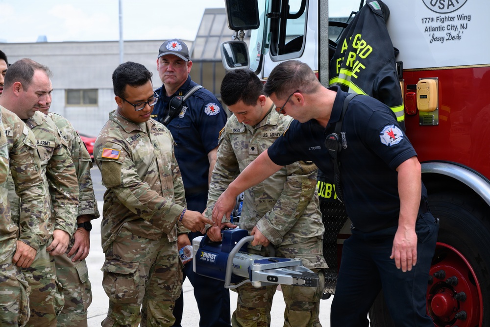 U.S. Army soldiers in OCP camouflage uniforms are receiving a demonstration of a Hurst rescue tool (commonly known as the 'Jaws of Life') from firefighters near a fire truck. The setting appears to be at the 177th Fighter Wing base in Atlantic City, NJ, suggesting an inter-service training or familiarization exercise. A civilian firefighter wearing a name tag reading 'Torres' is showing the equipment to the military personnel.