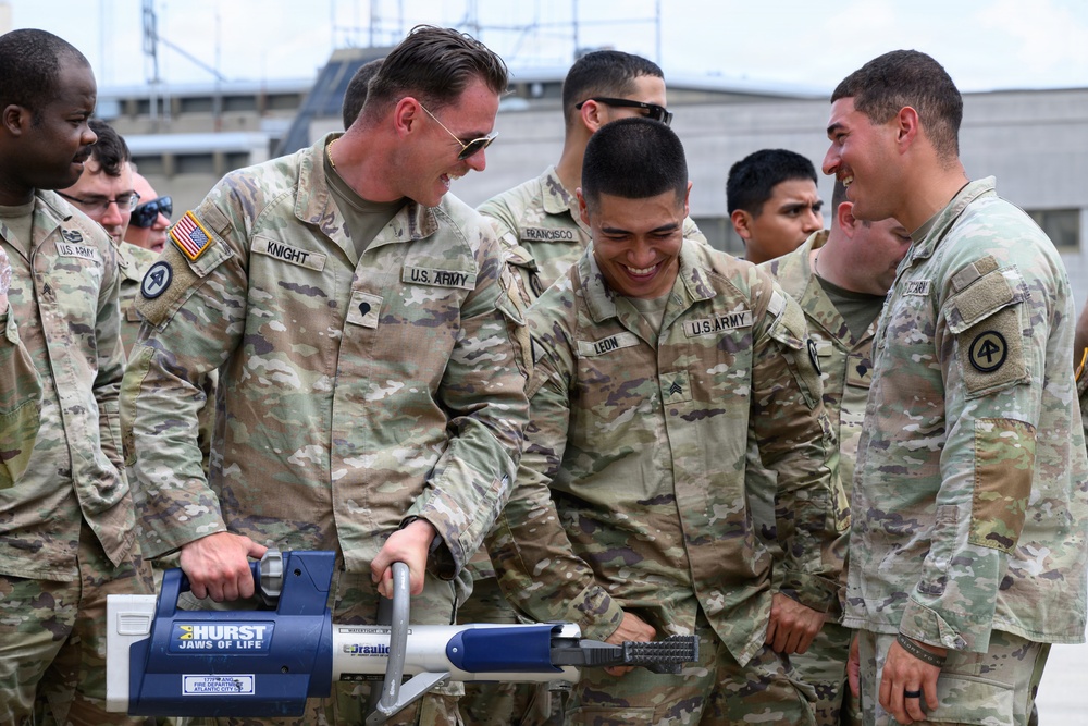 A group of U.S. Army soldiers in OCP (Operational Camouflage Pattern) uniforms are gathered together, laughing and smiling while examining a Hurst Jaws of Life hydraulic rescue tool. The mood is lighthearted and informal, suggesting a training demonstration or familiarization exercise. Name tapes visible include 'KNIGHT,' 'LEON,' and 'FRANCISCO,' all bearing U.S. ARMY identification.