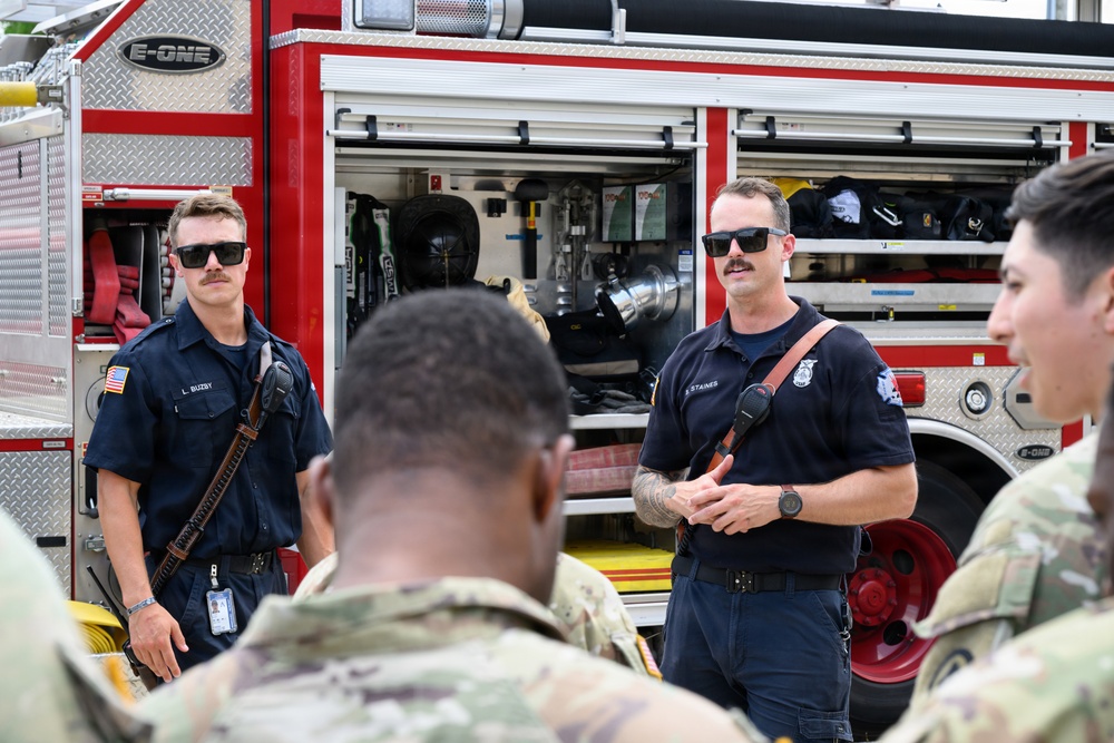 Two firefighters in navy blue uniforms are conducting a briefing or demonstration in front of a red E-ONE fire truck. They are addressing a group of military personnel in camouflage uniforms. The setting appears to be an outdoor training or orientation event at a military installation.