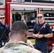 Two firefighters in navy blue uniforms are conducting a briefing or demonstration in front of a red E-ONE fire truck. They are addressing a group of military personnel in camouflage uniforms. The setting appears to be an outdoor training or orientation event at a military installation.