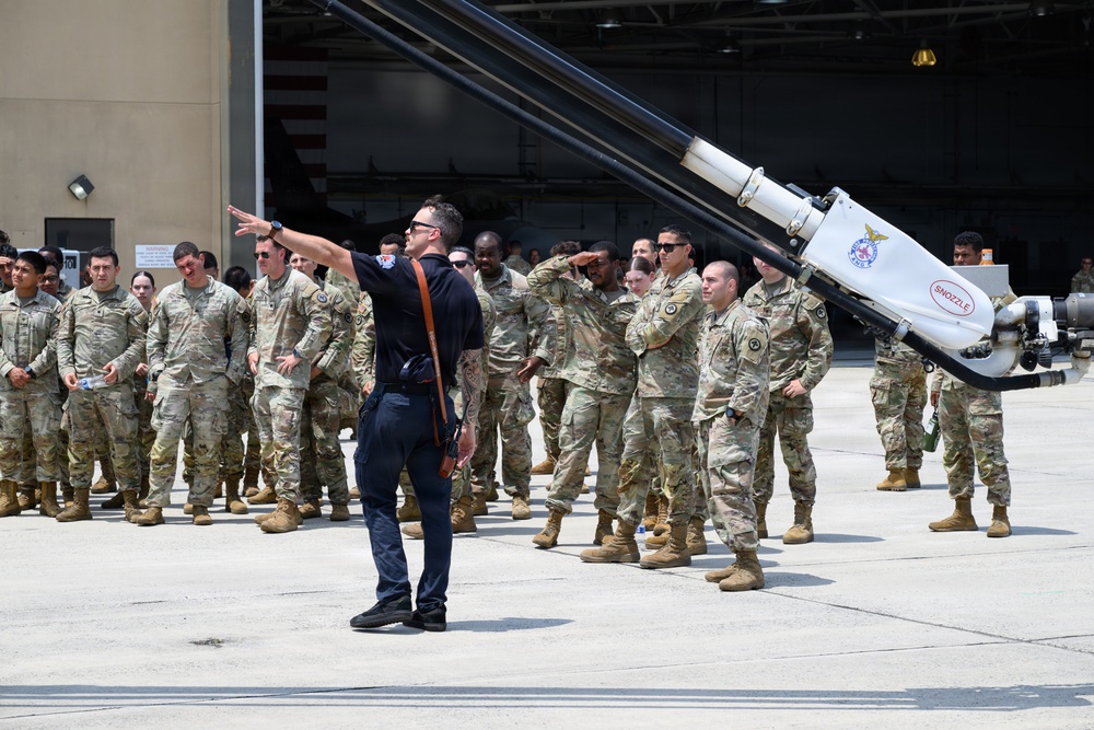 A civilian instructor or briefer in dark clothing is addressing a large group of military personnel in camouflage uniforms on an airfield tarmac. The group appears to be receiving a demonstration or orientation near a large piece of equipment labeled 'SNOZZLE', which appears to be an aircraft rescue firefighting (ARFF) apparatus. The setting is in front of a military hangar with an American flag visible in the background.