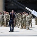 A civilian instructor or briefer in dark clothing is addressing a large group of military personnel in camouflage uniforms on an airfield tarmac. The group appears to be receiving a demonstration or orientation near a large piece of equipment labeled 'SNOZZLE', which appears to be an aircraft rescue firefighting (ARFF) apparatus. The setting is in front of a military hangar with an American flag visible in the background.