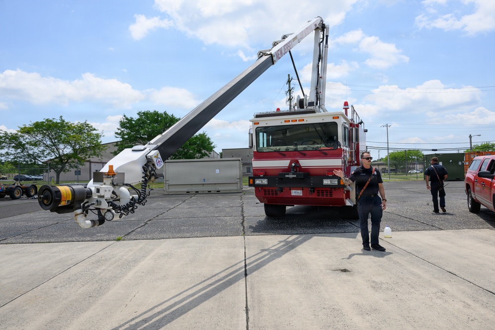 A red fire truck equipped with a specialized articulating boom arm or crane is parked on a concrete lot, with the boom extended outward. Two personnel in dark uniforms are present — one appears to be demonstrating or explaining the equipment. The setting appears to be an outdoor training or demonstration area, possibly affiliated with New Jersey based on text visible on the boom.