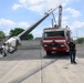 A red fire truck equipped with a specialized articulating boom arm or crane is parked on a concrete lot, with the boom extended outward. Two personnel in dark uniforms are present — one appears to be demonstrating or explaining the equipment. The setting appears to be an outdoor training or demonstration area, possibly affiliated with New Jersey based on text visible on the boom.