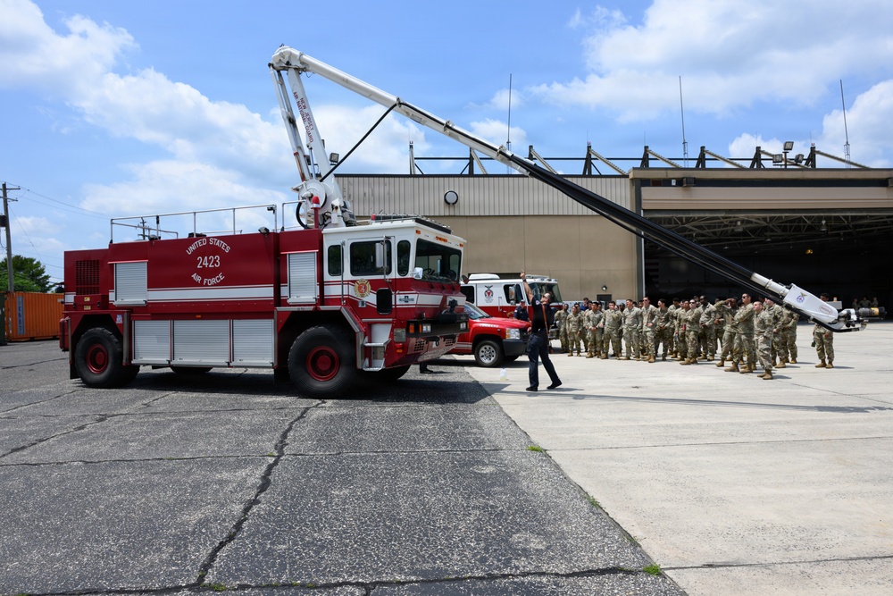 A United States Air Force fire truck (numbered 2423) is displayed on a tarmac apron in front of a large hangar facility. A group of military personnel in camouflage uniforms are assembled in formation to the right, appearing to observe a demonstration or ceremony. One individual in dark clothing stands between the truck and the formation, possibly addressing the group.