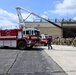 A United States Air Force fire truck (numbered 2423) is displayed on a tarmac apron in front of a large hangar facility. A group of military personnel in camouflage uniforms are assembled in formation to the right, appearing to observe a demonstration or ceremony. One individual in dark clothing stands between the truck and the formation, possibly addressing the group.