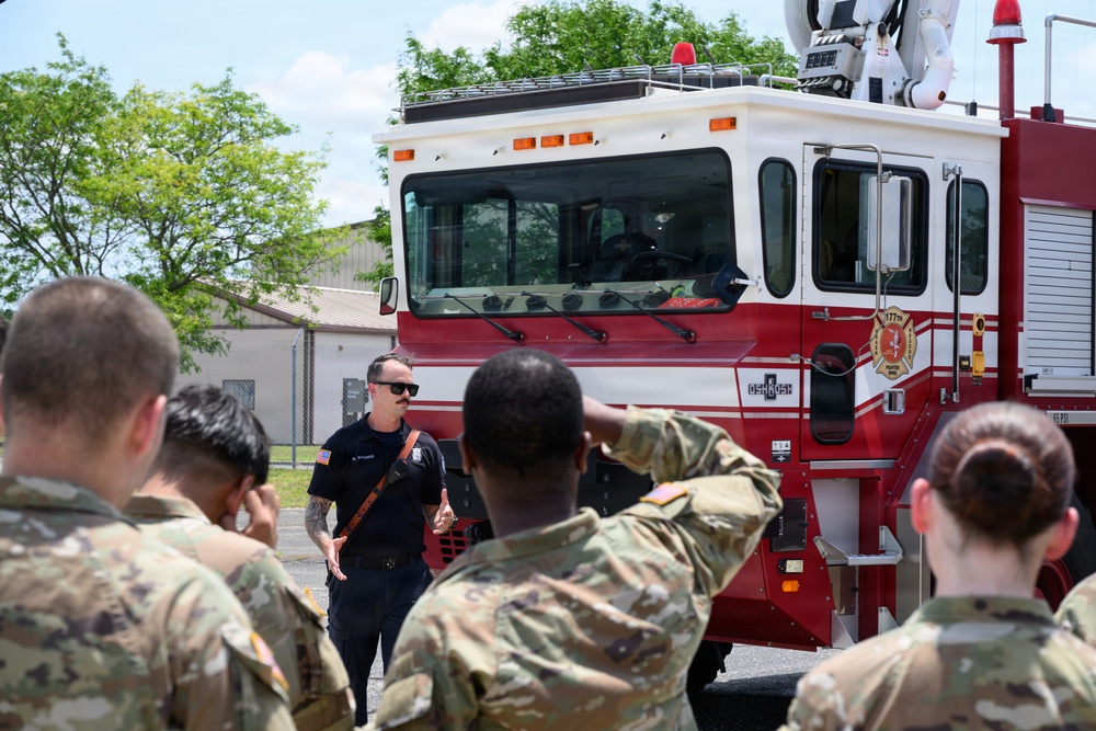 A civilian firefighter or fire department instructor is briefing a group of military personnel in camouflage uniforms in front of a red and white fire truck. The setting appears to be an outdoor area on a military installation, suggesting a training or familiarization exercise. The firefighter is gesturing and speaking while the soldiers attentively face him.