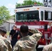 A civilian firefighter or fire department instructor is briefing a group of military personnel in camouflage uniforms in front of a red and white fire truck. The setting appears to be an outdoor area on a military installation, suggesting a training or familiarization exercise. The firefighter is gesturing and speaking while the soldiers attentively face him.