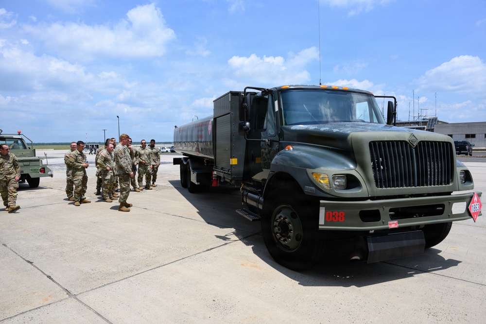 A group of military personnel in camouflage uniforms are gathered on an airfield tarmac beside a large olive-drab fuel tanker truck numbered '038'. The scene appears to be a training, inspection, or briefing event related to aircraft refueling operations. The setting is consistent with a military air base, with airport infrastructure visible in the background.