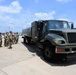 A group of military personnel in camouflage uniforms are gathered on an airfield tarmac beside a large olive-drab fuel tanker truck numbered '038'. The scene appears to be a training, inspection, or briefing event related to aircraft refueling operations. The setting is consistent with a military air base, with airport infrastructure visible in the background.