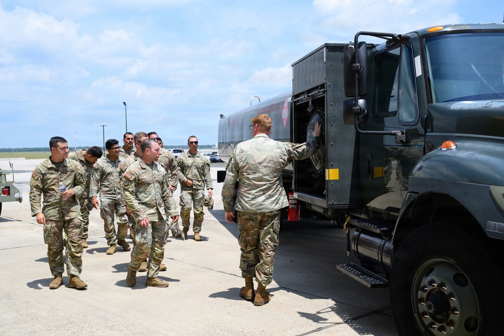 A group of military personnel in OCP (Operational Camouflage Pattern) uniforms are gathered on what appears to be an airfield tarmac. They are examining or receiving a briefing near a large military fuel tanker truck. The setting suggests a training exercise, orientation, or operational briefing related to aircraft refueling operations.
