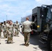 A group of military personnel in OCP (Operational Camouflage Pattern) uniforms are gathered on what appears to be an airfield tarmac. They are examining or receiving a briefing near a large military fuel tanker truck. The setting suggests a training exercise, orientation, or operational briefing related to aircraft refueling operations.