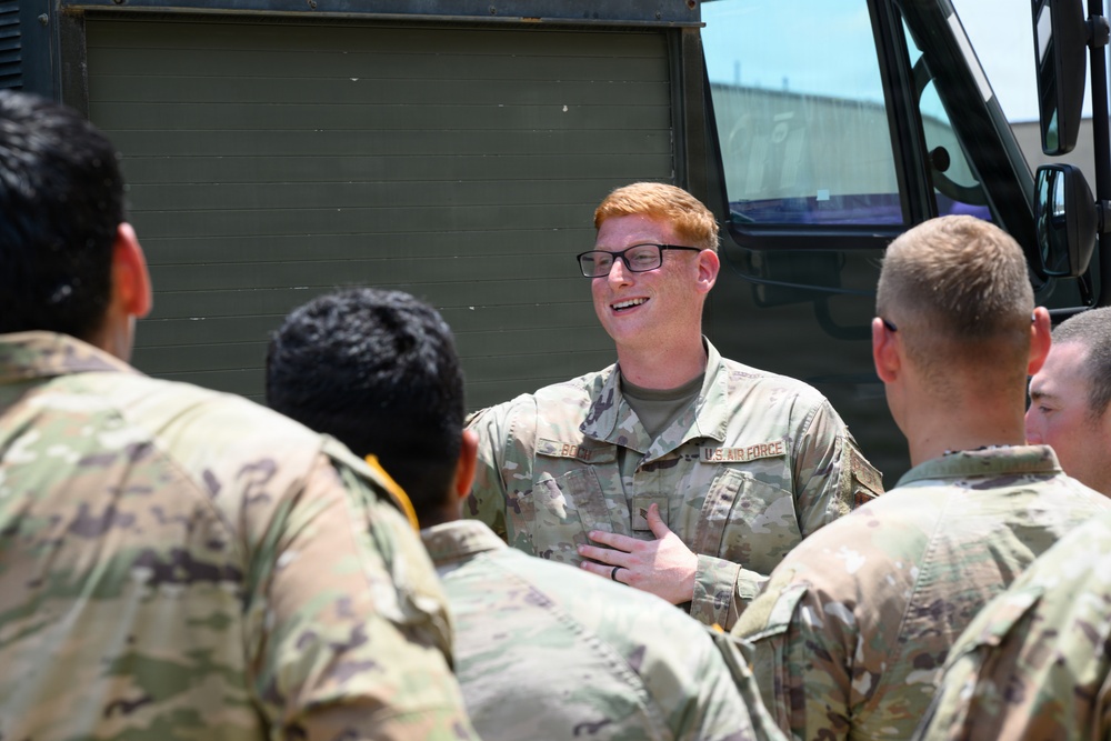 A U.S. Air Force service member with red hair and glasses is speaking or briefing a small group of fellow service members in camouflage uniforms outdoors. The central figure appears engaged and smiling, with one hand placed on his chest, suggesting an informal or instructional conversation. A large military vehicle is visible in the background.