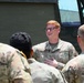 A U.S. Air Force service member with red hair and glasses is speaking or briefing a small group of fellow service members in camouflage uniforms outdoors. The central figure appears engaged and smiling, with one hand placed on his chest, suggesting an informal or instructional conversation. A large military vehicle is visible in the background.