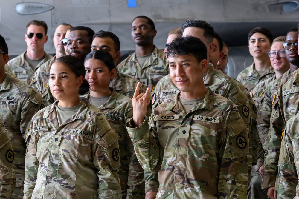 A group of U.S. Army soldiers in OCP (Operational Camouflage Pattern) uniforms are assembled together, likely in a hangar or large military facility. The soldiers display a diverse composition and are standing in formation or a group pose. One soldier in the foreground, identified by the name tape 'CARLOS,' is raising his hand with two fingers extended.