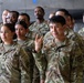 A group of U.S. Army soldiers in OCP (Operational Camouflage Pattern) uniforms are assembled together, likely in a hangar or large military facility. The soldiers display a diverse composition and are standing in formation or a group pose. One soldier in the foreground, identified by the name tape 'CARLOS,' is raising his hand with two fingers extended.