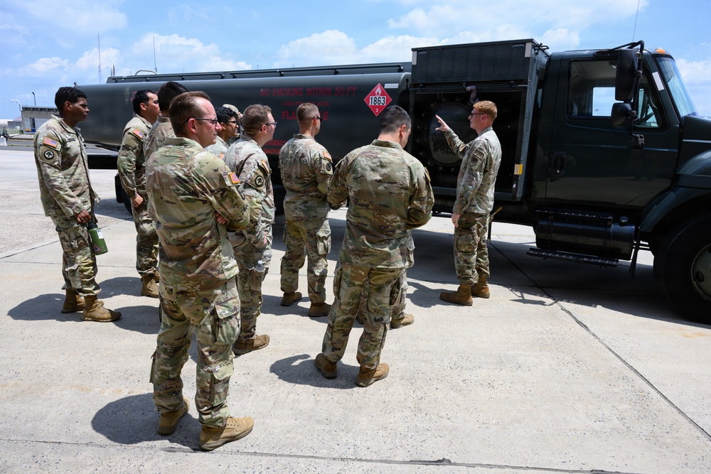 A group of military personnel in camouflage uniforms are gathered around a large fuel tanker truck on what appears to be a military installation or airfield. One soldier appears to be conducting a briefing or demonstration, pointing at the truck's equipment panel. The tanker displays hazmat placard number 1863, indicating aviation fuel.