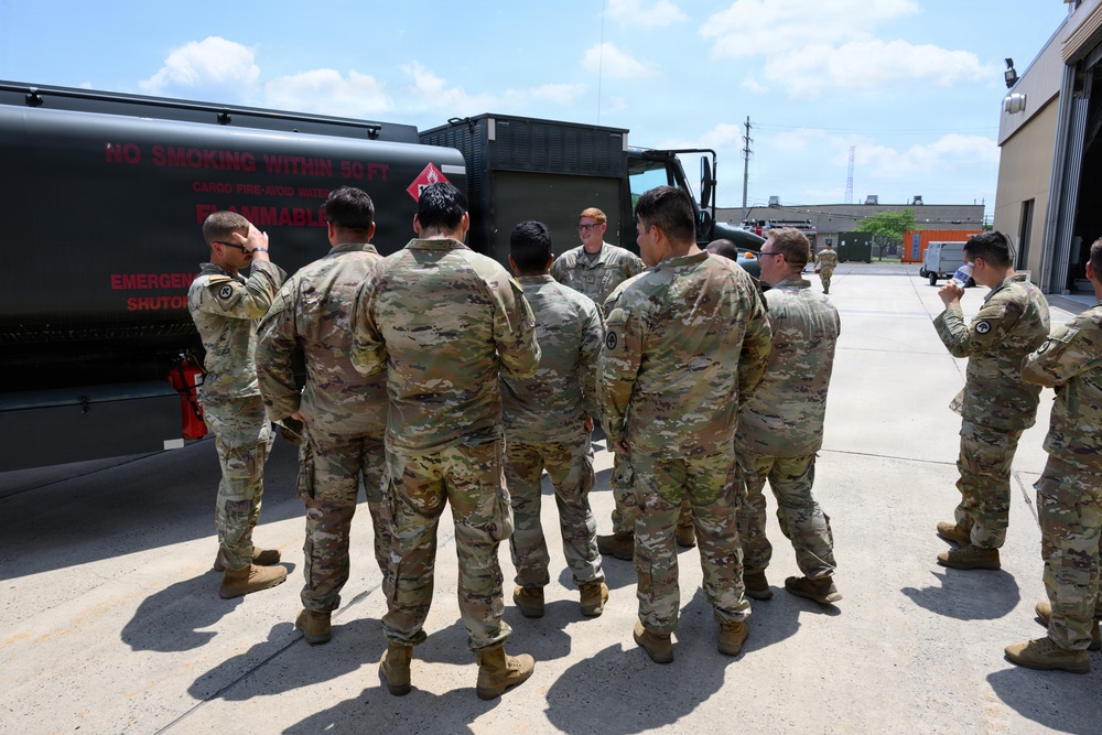 A group of military personnel in OCP (Operational Camouflage Pattern) uniforms are gathered around a fuel tanker truck for what appears to be a training or briefing session. One soldier facing the group appears to be instructing or addressing the others near the tanker. The setting is an outdoor military installation or motor pool area.