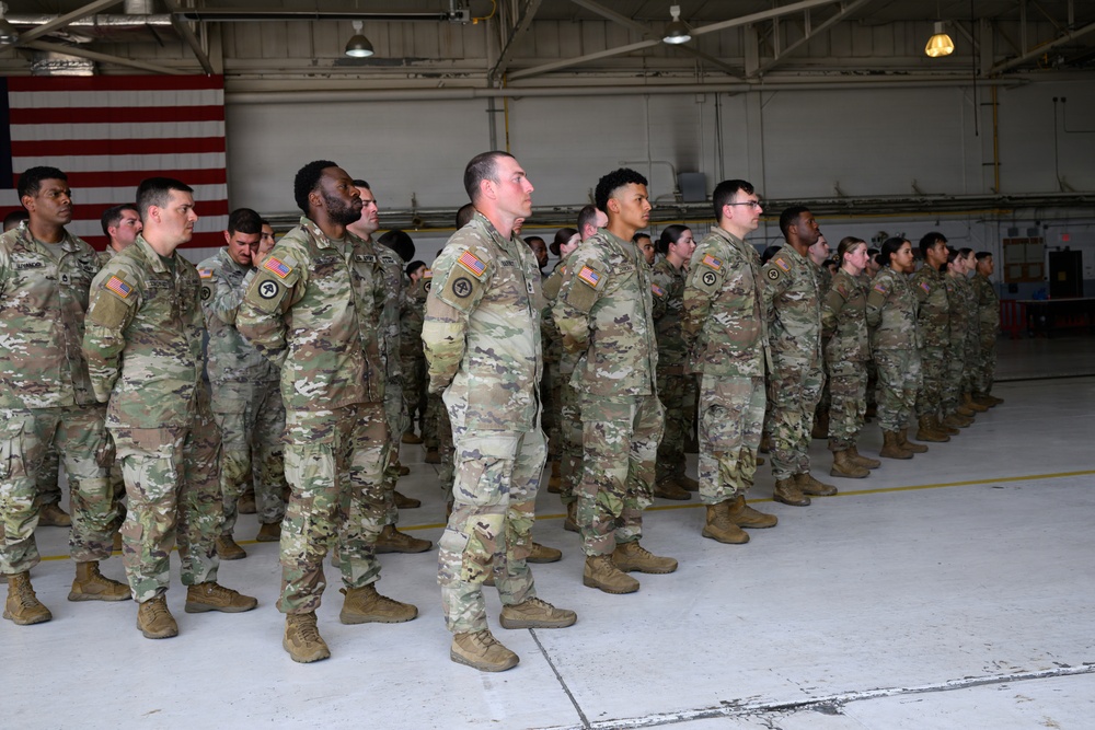 A formation of U.S. Army soldiers standing at attention inside a military hangar. The soldiers are wearing Operational Camouflage Pattern (OCP) uniforms with American flag patches visible on their right shoulders. The setting appears to be a formal military ceremony or unit formation event.
