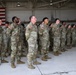 A formation of U.S. Army soldiers standing at attention inside a military hangar. The soldiers are wearing Operational Camouflage Pattern (OCP) uniforms with American flag patches visible on their right shoulders. The setting appears to be a formal military ceremony or unit formation event.