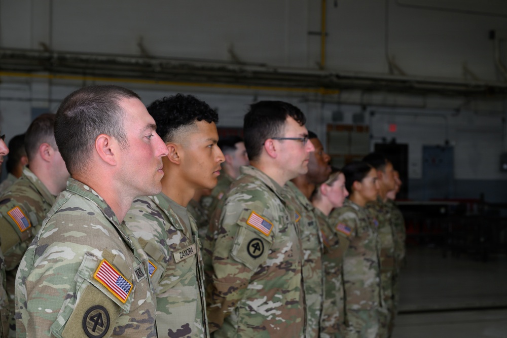 A group of U.S. Army soldiers in Operational Camouflage Pattern (OCP) uniforms are standing at attention in what appears to be a military hangar or large indoor facility. The soldiers are arranged in formation, facing the same direction in profile view. American flag patches and unit insignia are visible on their uniforms.