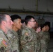 A group of U.S. Army soldiers in Operational Camouflage Pattern (OCP) uniforms are standing at attention in what appears to be a military hangar or large indoor facility. The soldiers are arranged in formation, facing the same direction in profile view. American flag patches and unit insignia are visible on their uniforms.