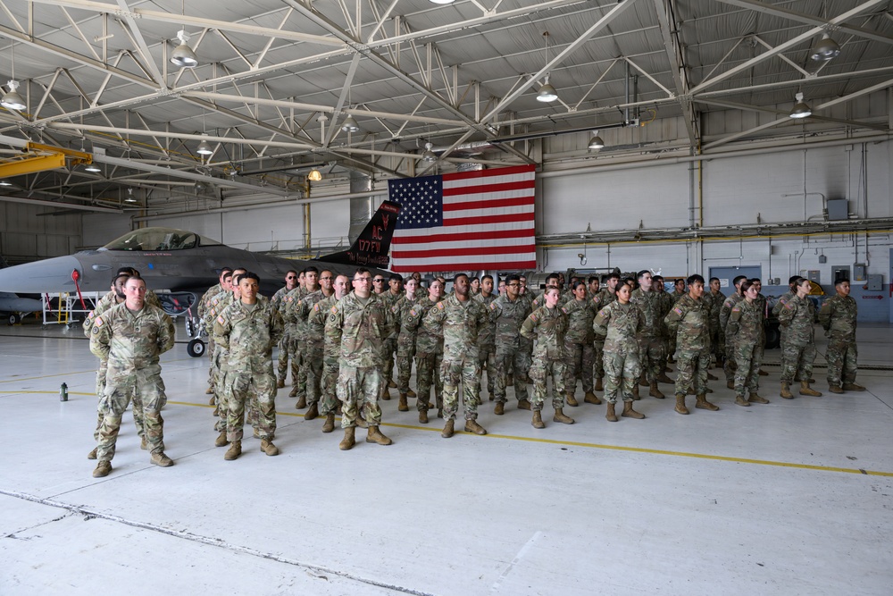 A large group of military personnel in camouflage uniforms are assembled in formation inside an aircraft hangar. Behind them stands a fighter jet (appearing to be an F-16) and a large American flag draped on the hangar wall. The gathering appears to be a formal military ceremony or formation event.