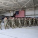 A large group of military personnel in camouflage uniforms are assembled in formation inside an aircraft hangar. Behind them stands a fighter jet (appearing to be an F-16) and a large American flag draped on the hangar wall. The gathering appears to be a formal military ceremony or formation event.