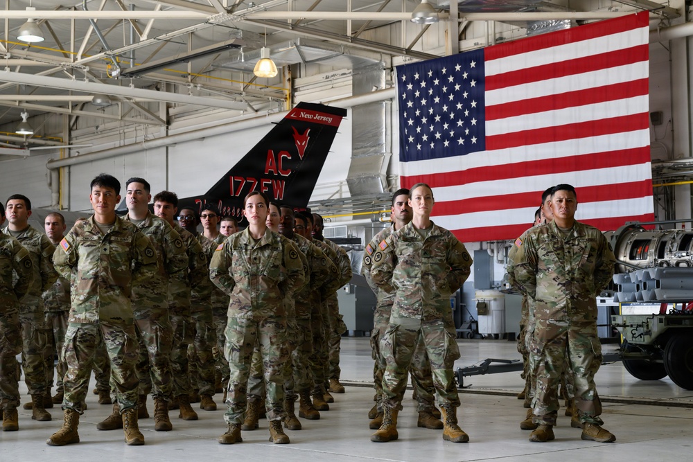 A formation of U.S. military personnel in OCP (Operational Camouflage Pattern) uniforms stand at parade rest inside a military hangar. Behind them is a large American flag and the tail section of a military aircraft bearing the markings 'AC 177FW' and 'New Jersey', identifying this as the 177th Fighter Wing of the New Jersey Air National Guard. The setting appears to be a formal military ceremony or briefing.