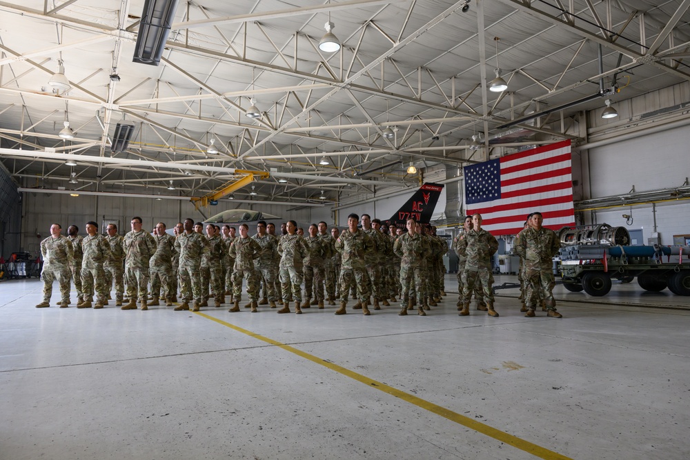 A large group of military personnel in camouflage uniforms are standing in formation inside an aircraft hangar. An American flag is prominently displayed in the background alongside a military aircraft with tail markings 'AC 177FW'. The setting appears to be an official military ceremony or formation event.