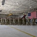 A large group of military personnel in camouflage uniforms are standing in formation inside an aircraft hangar. An American flag is prominently displayed in the background alongside a military aircraft with tail markings 'AC 177FW'. The setting appears to be an official military ceremony or formation event.