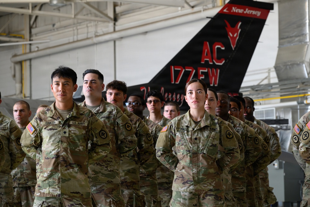 A group of U.S. Army soldiers in OCP (Operational Camouflage Pattern) uniforms stand at parade rest inside a military hangar. Behind them is the tail section of a black aircraft bearing the markings 'AC 177 FW' and 'New Jersey,' identifying it as belonging to the 177th Fighter Wing of the New Jersey Air National Guard. Two soldiers in the foreground are identifiable by name tapes: 'CARLOS' and 'PAEZ.'