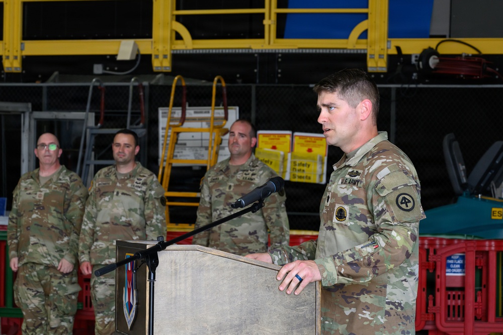 A U.S. Army soldier is delivering a speech at a podium during what appears to be a military ceremony or briefing held inside an industrial or hangar facility. Three other soldiers in matching OCP (Operational Camouflage Pattern) uniforms stand at attention in the background. A military unit insignia is visible on the front of the podium.