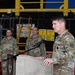 A U.S. Army soldier is delivering a speech at a podium during what appears to be a military ceremony or briefing held inside an industrial or hangar facility. Three other soldiers in matching OCP (Operational Camouflage Pattern) uniforms stand at attention in the background. A military unit insignia is visible on the front of the podium.