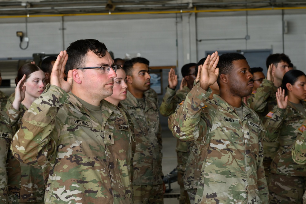 A group of U.S. Army soldiers are raising their right hands in what appears to be a re-enlistment or oath of enlistment ceremony. The soldiers are wearing Army Combat Uniforms (ACU) in Operational Camouflage Pattern (OCP). The ceremony is taking place indoors, likely in a military hangar or large facility.