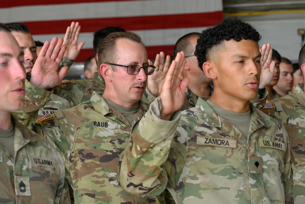 A group of U.S. Army soldiers are raising their right hands in what appears to be a military oath ceremony, likely a reenlistment or enlistment swearing-in. The soldiers are wearing OCP (Operational Camouflage Pattern) uniforms with name tapes visible, including 'RAUB' and 'ZAMORA'. An American flag is partially visible in the background, consistent with a formal military ceremony setting.