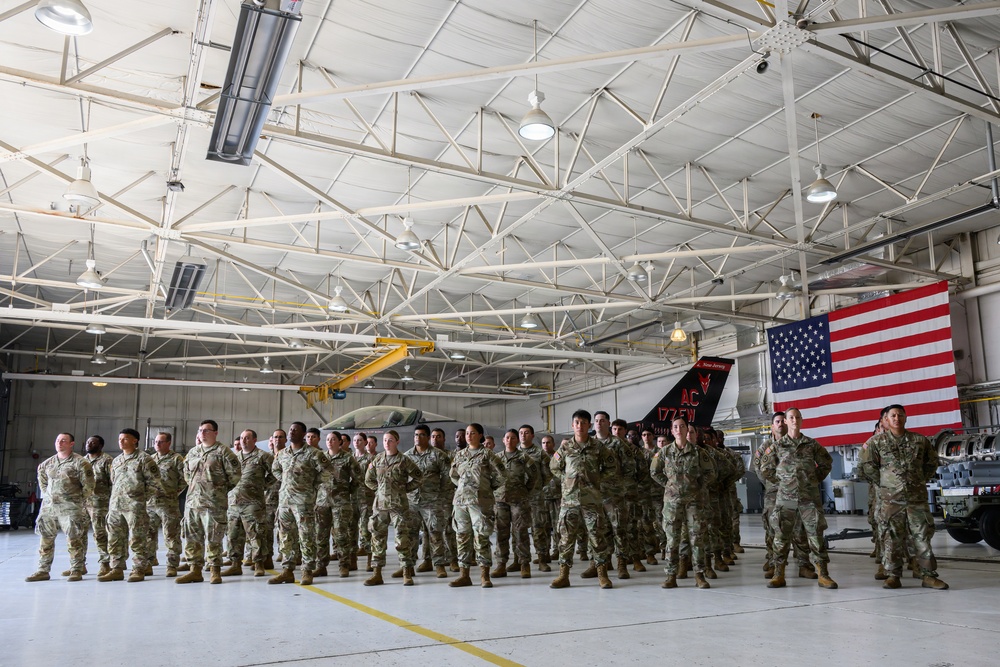 A large formation of military personnel in camouflage uniforms (OCP pattern) stands at attention inside a military aircraft hangar. An American flag is prominently displayed in the background alongside what appears to be a military jet aircraft with tail markings reading 'AC 177FW'. The setting suggests a formal military ceremony or briefing event.
