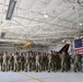 A large formation of military personnel in camouflage uniforms (OCP pattern) stands at attention inside a military aircraft hangar. An American flag is prominently displayed in the background alongside what appears to be a military jet aircraft with tail markings reading 'AC 177FW'. The setting suggests a formal military ceremony or briefing event.