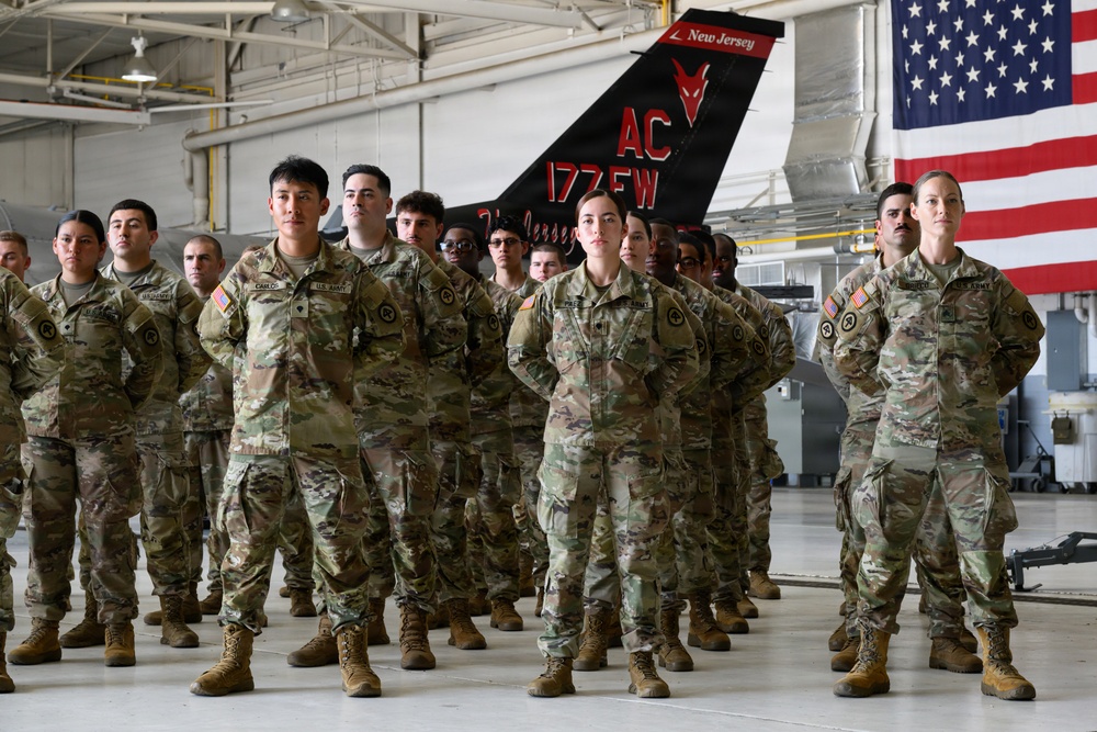 A formation of U.S. Army soldiers in MultiCam/OCP camouflage uniforms standing at parade rest inside a military hangar. Behind them is a fighter jet tail fin marked 'New Jersey AC 177 FW' (177th Fighter Wing) and a large American flag. The setting appears to be a formal military ceremony or briefing at what is likely Atlantic City Air National Guard Base.