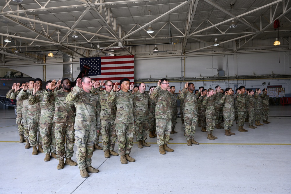 A large group of military personnel in OCP (Operational Camouflage Pattern) uniforms are raising their right hands in what appears to be a formal oath ceremony inside a military hangar. An American flag is prominently displayed on the back wall, serving as a ceremonial backdrop. This appears to be a reenlistment or enlistment ceremony conducted in a military aviation hangar.