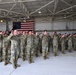 A large group of military personnel in OCP (Operational Camouflage Pattern) uniforms are raising their right hands in what appears to be a formal oath ceremony inside a military hangar. An American flag is prominently displayed on the back wall, serving as a ceremonial backdrop. This appears to be a reenlistment or enlistment ceremony conducted in a military aviation hangar.