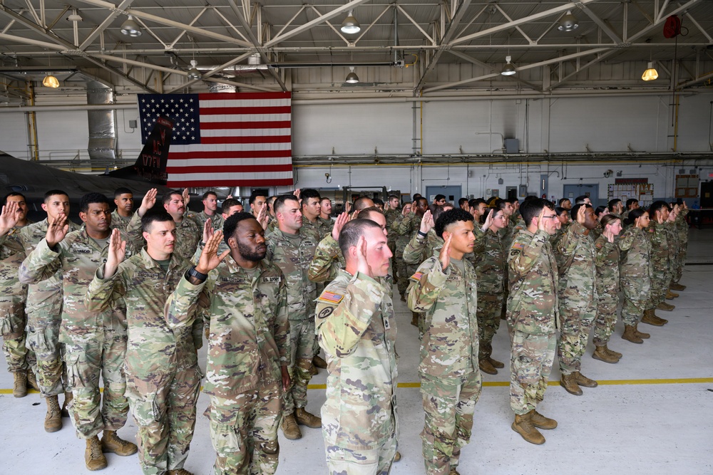 A large group of U.S. Army soldiers in OCP (Operational Camouflage Pattern) uniforms are raising their right hands in what appears to be a military oath ceremony, likely a reenlistment or naturalization ceremony. The event is taking place inside a military hangar with an American flag prominently displayed on the back wall. The formation consists of dozens of soldiers standing in organized rows.