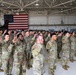 A large group of U.S. Army soldiers in OCP (Operational Camouflage Pattern) uniforms are raising their right hands in what appears to be a military oath ceremony, likely a reenlistment or naturalization ceremony. The event is taking place inside a military hangar with an American flag prominently displayed on the back wall. The formation consists of dozens of soldiers standing in organized rows.