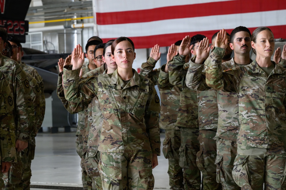 A group of U.S. Army soldiers in OCP (Operational Camouflage Pattern) uniforms are raising their right hands in what appears to be a military oath ceremony, likely a reenlistment or enlistment swearing-in. The ceremony is taking place inside a military hangar with a large American flag displayed in the background. Multiple soldiers are visible in formation, with name tapes and U.S. Army patches clearly identifiable.