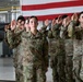 A group of U.S. Army soldiers in OCP (Operational Camouflage Pattern) uniforms are raising their right hands in what appears to be a military oath ceremony, likely a reenlistment or enlistment swearing-in. The ceremony is taking place inside a military hangar with a large American flag displayed in the background. Multiple soldiers are visible in formation, with name tapes and U.S. Army patches clearly identifiable.