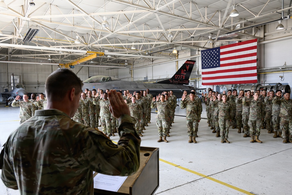 A military re-enlistment or oath of office ceremony is taking place inside an aircraft hangar. A uniformed officer stands at a podium administering an oath to a large group of service members who have their right hands raised. A large American flag and a military jet aircraft are prominently displayed in the background.