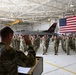 A military re-enlistment or oath of office ceremony is taking place inside an aircraft hangar. A uniformed officer stands at a podium administering an oath to a large group of service members who have their right hands raised. A large American flag and a military jet aircraft are prominently displayed in the background.